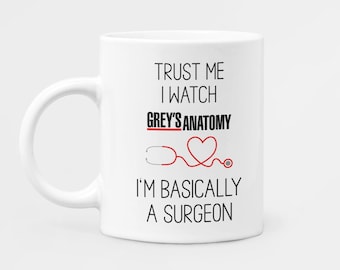 May include: White ceramic mug with black text that reads "Trust me I watch Grey's Anatomy I'm basically a surgeon". A red heart and stethoscope are illustrated on the mug.
