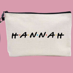 May include: White canvas zippered pouch with a black zipper and a black strap. The pouch has the name "HANNAH" written in black letters with colourful dots between each letter.
