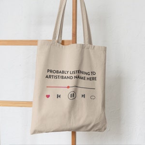 May include: A beige tote bag featuring the text "PROBABLY LISTENING TO ARTIST/BAND NAME HERE" in black. Below the text is a music player graphic with a pink heart, rewind, pause, fast forward, and repeat icons.