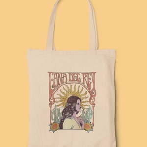 May include: A white canvas tote bag with a vintage-style graphic of Lana Del Rey. The graphic features a woman with long dark hair, a sun, and cacti. The text "Lana Del Rey" is in a retro font.
