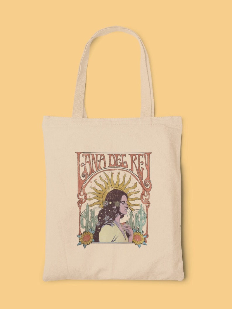 May include: A white canvas tote bag with a vintage-style graphic of Lana Del Rey. The graphic features a woman with long dark hair, a sun, and cacti. The text "Lana Del Rey" is in a retro font.