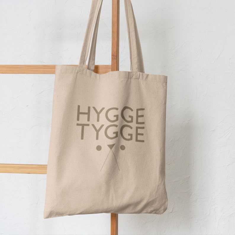 May include: A beige canvas tote bag with the text "HYGGE TYGGE" printed in brown. The bag has two brown dots and a brown X shape below the text.