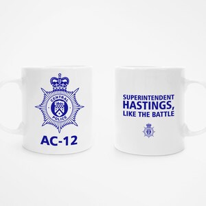 May include: White ceramic mug with a blue police badge and the text "AC-12" on one side and the text "Superintendent Hastings, Like the Battle" on the other side.
