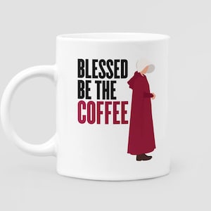 May include: White ceramic coffee mug with the text "BLESSED BE THE COFFEE" in black and burgundy. A burgundy-robed figure with a white bonnet is on the right side of the mug. The mug has a white handle.