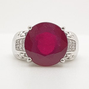 May include: A silver ring with a large, oval-shaped ruby gemstone surrounded by smaller, clear gemstones.