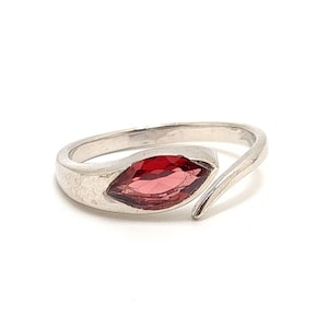 May include: A silver ring with a red marquise-shaped gemstone set in the center. The band is thin and wraps around the finger.