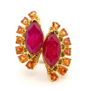 May include: A gold-toned ring featuring two large, oval-cut, deep red gemstones. The ring is adorned with smaller orange and green accent stones, creating a symmetrical, butterfly-like design. The ring is set against a white background.