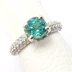 Puede incluir: Un anillo de plata con una gran piedra preciosa redonda de color verde azulado rodeada de pequeñas piedras preciosas blancas. El anillo está colocado sobre un fondo blanco.