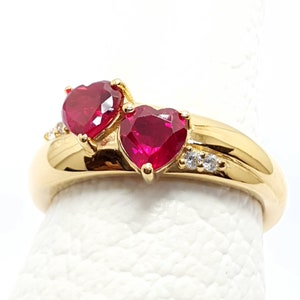 Puede incluir: Un anillo de oro con dos piedras preciosas de rubí en forma de corazón y piedras preciosas más pequeñas de color claro engastadas en la banda.