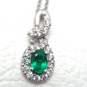 Puede incluir: Un collar de plata con un colgante que tiene una esmeralda verde rodeada de piedras preciosas transparentes más pequeñas. El colgante tiene la forma de un nudo estilizado.