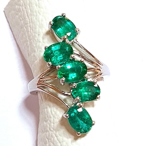 Puede incluir: Un anillo de plata con un grupo de cinco piedras preciosas de color verde esmeralda de forma ovalada. El anillo tiene un diseño delicado y curvo.