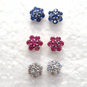 以下が含まれることがあります： 3組の花型のスタッドピアス。 上のペアは濃い青色の宝石、中央のペアは鮮やかな赤い宝石、下のペアは透明で輝く石が特徴です。 すべて銀色の金属にセットされています。