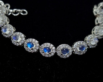 Puede incluir: Una pulsera de plata con una fila de piedras de luna azul ovaladas rodeadas de piedras transparentes más pequeñas.