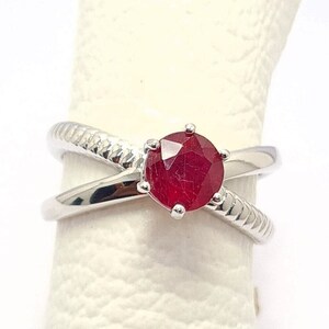 May include: A silver ring with a red gemstone set in the centre. The ring has a criss-cross design.