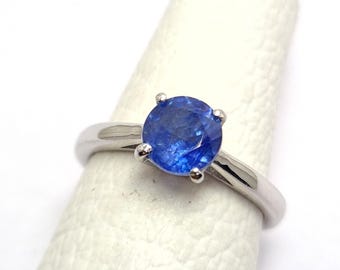 Op de afbeelding: Een zilveren ring met een grote, ronde, briljant geslepen blauwe edelsteen. De edelsteen wordt op zijn plaats gehouden door vier pootjes. De ring wordt getoond op een witte ondergrond, waarbij het ontwerp en de kleur van de edelsteen worden benadrukt.