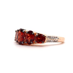May include: A rose gold ring featuring three prominent oval-cut deep red garnet gemstones set in a row, flanked by smaller round-cut garnets, with a band accented by a channel of tiny clear white stones along the sides of the metal shank.