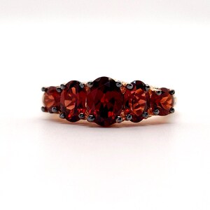 May include: A ring featuring five oval-cut deep red garnet gemstones arranged in a horizontal row. The stones are set in a dark metal prong setting that secures each individual garnet, creating a continuous line of faceted red gems across the band.