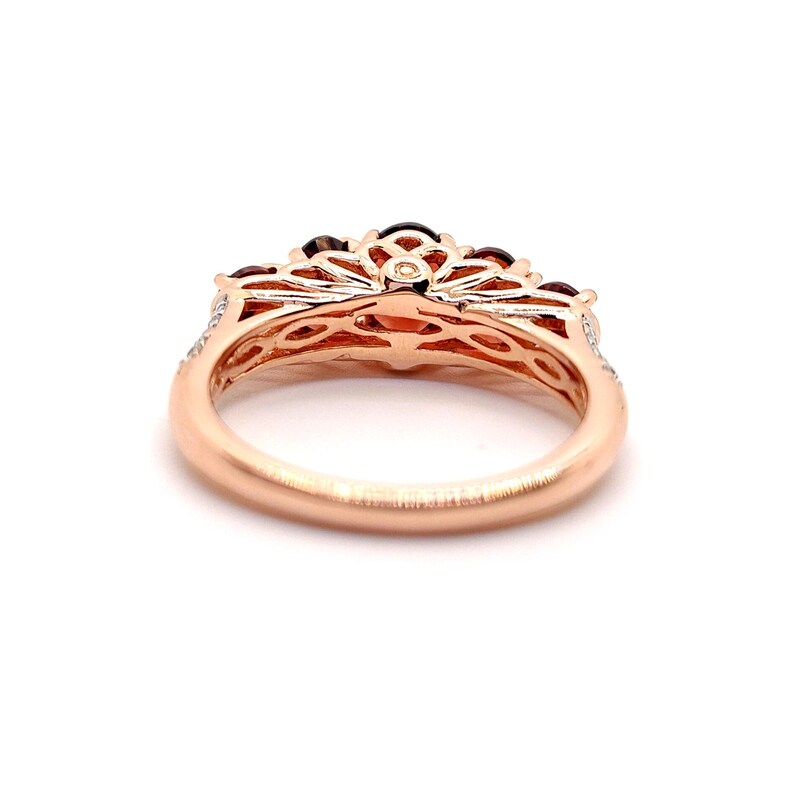 May include: A rose gold ring featuring a band with a smooth finish and an intricate, openwork gallery design. The setting holds multiple dark red, round-cut gemstones arranged in a fan-like pattern across the top of the metal structure.