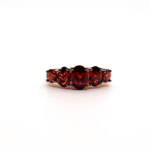 May include: A ring featuring five oval-cut deep red garnet gemstones arranged in a row. The stones are set in a dark metal prong setting that secures each individual gem, creating a continuous horizontal line of faceted red stones across the band.