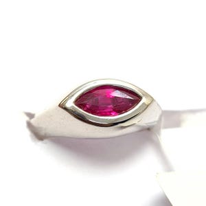 Könnte beinhalten: Ein silberner Ring mit einem marquiseförmigen, leuchtend roten Edelstein. Der Edelstein ist in einer Lünettenfassung gefasst, wodurch ein schlankes Design entsteht. Der Ring wird vor weißem Hintergrund gezeigt, um die polierte Oberfläche hervorzuheben.