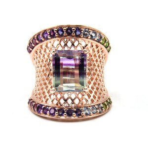 May include: A rose gold ring with a large, square-shaped, multi-colored gemstone set in a delicate, openwork design. The ring is accented with a row of small, colorful gemstones around the band.
