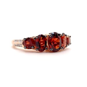 May include: A rose gold ring featuring five oval-cut deep red garnet gemstones arranged in a row. The center stone is the largest, flanked by two smaller garnets on each side. The band is accented with small clear stones along the side shank.