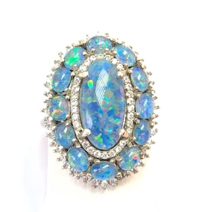 May include: An oval ring with a large, iridescent blue opal at its centre, encircled by smaller blue opals and clear gemstones. The ring has a silver-coloured metal setting, creating a luxurious and eye-catching design.
