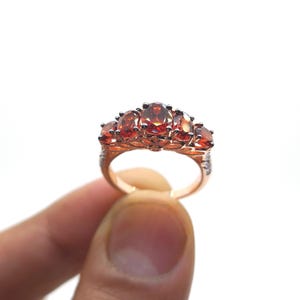 May include: A rose gold ring featuring five oval-cut, deep orange-red gemstones arranged in a row. The metal band displays intricate filigree detailing beneath the stones, and the sides of the band are accented with small, clear, sparkling stones.