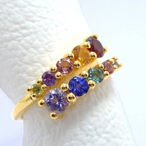 May include: A gold ring with a rainbow of gemstones. The ring is set with a row of round, faceted stones in shades of green, purple, blue, pink, orange, and red.