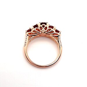 May include: A rose gold ring featuring a series of deep red, round-cut gemstones set along the top. The metal band displays an intricate, openwork filigree design with small, clear accent stones embedded along the sides of the shank.