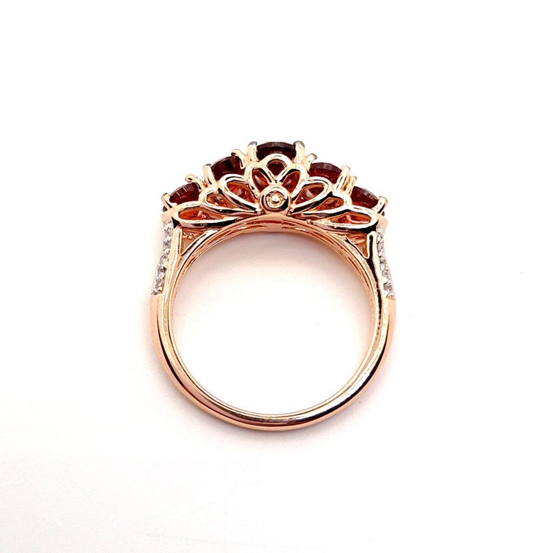 May include: A rose gold ring featuring a series of deep red, round-cut gemstones set along the top. The metal band displays an intricate, openwork filigree design with small, clear accent stones embedded along the sides of the shank.