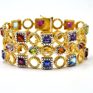 May include: A gold bracelet with a multi-colored gemstone design. The bracelet features a series of round and square gemstones in various colors, including red, blue, green, purple, and orange.