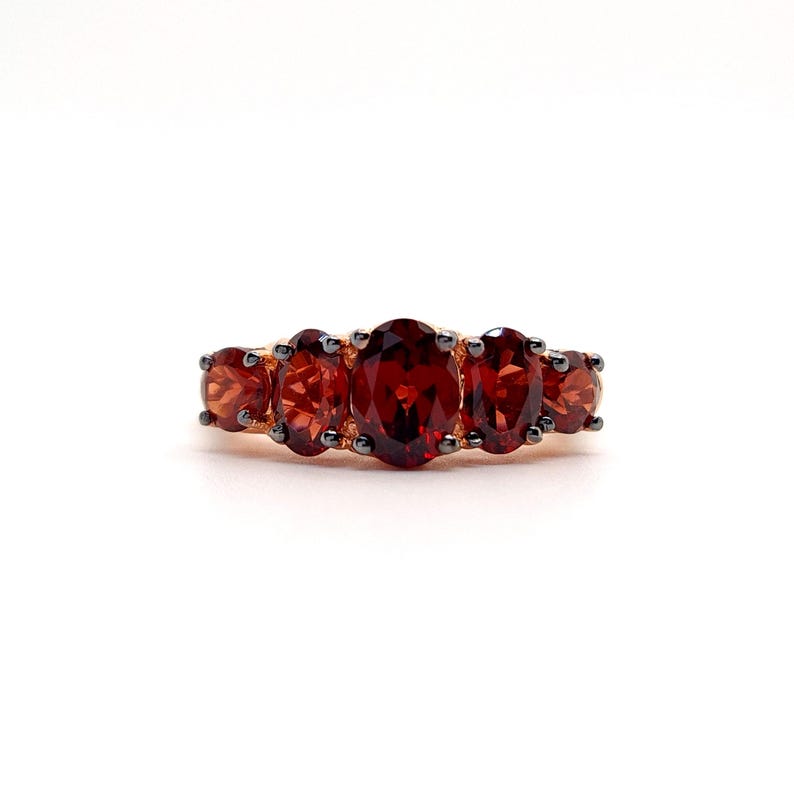May include: A ring featuring five oval-cut deep red garnet gemstones set in a dark metal band. The center stone is the largest, flanked by two smaller stones on each side, all secured with dark prongs that contrast with the vibrant red color.