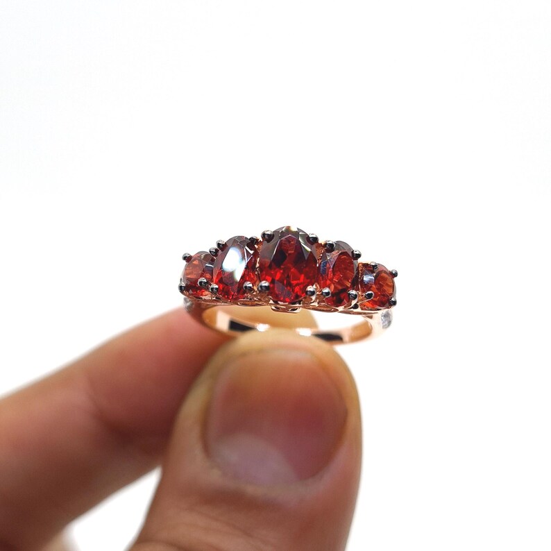 May include: A ring featuring five oval-cut deep red gemstones set in a polished metal band. The central stone is the largest, flanked by two smaller stones on each side, all secured with metal prongs in a horizontal arrangement across the finger.