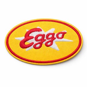 May include: Embroidered patch featuring the word "Eggo" in red, set against a white starburst on a yellow oval. The oval has a red border. The patch is designed for application on clothing or accessories.
