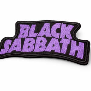 May include: Embroidered patch with the words "BLACK SABBATH" in purple, bold lettering. The patch has a black border and backing, with a curved shape. Suitable for application on clothing or accessories.