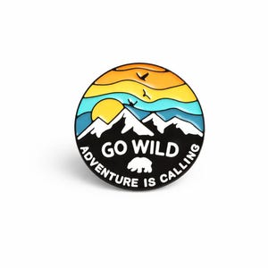 Può includere: Spilla rotonda in smalto con un design di montagna e tramonto. La spilla ha un bordo nero con le parole "GO WILD" e "ADVENTURE IS CALLING" in bianco. Una silhouette di orso è al centro.