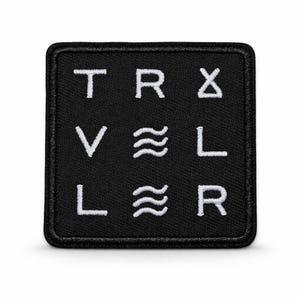 Könnte beinhalten: Schwarzer quadratischer Aufnäher mit weiß gesticktem Text und Symbolen. Das Wort „TRAVELER“ wird mit Symbolen geschrieben. Der Aufnäher hat einen schwarzen Rand und ist wahrscheinlich zum Aufnähen auf Kleidung oder Taschen bestimmt.