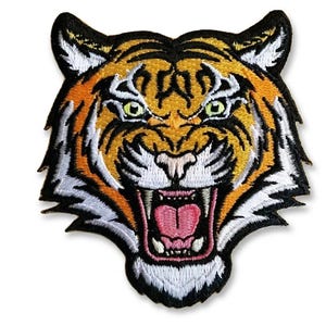 Könnte beinhalten: Gestickter Tigerkopf-Aufnäher mit orangefarbenen, schwarzen, weißen und gelben Details. Der Tiger brüllt, mit einer rosa Zunge und scharfen weißen Zähnen. Der Aufnäher hat einen schwarzen Rand.