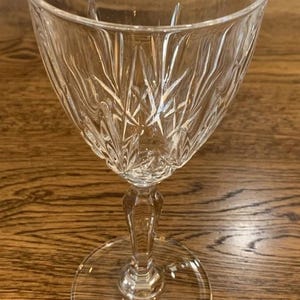 May include: A clear crystal wine glass with a detailed, etched pattern. The glass has a rounded bowl, a slender stem, and a circular base. The etched design features a series of vertical and diagonal lines, creating a starburst effect.