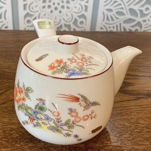 Könnte beinhalten: Weiße Teekanne aus Keramik mit floralem Muster und rotem Rand. Die Teekanne hat einen Deckel mit einem kleinen Knopf und einem Ausguss. Die Teekanne ist mit Vögeln und Blumen verziert.