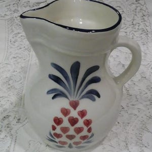 May include: White ceramic pitcher with a blue rim and handle. The pitcher features a hand-painted design of a pineapple made of red hearts and blue leaves. A decorative kitchenware item.