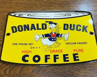 Może przedstawiać: Żółty metalowy znak z kreskówkowym wizerunkiem Donalda Kaczora trzymającego puszkę kawy. Na znaku widnieje napis "Donald Duck High Grade Pure Coffee" oraz "One Pound Net Vacuum Packed".