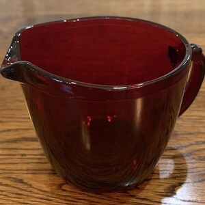 Peut inclure: Une cruche en verre rouge rubis avec une poignée et un bec verseur. La cruche est faite d'un matériau translucide, permettant à la lumière de passer à travers. La cruche est posée sur une surface en bois.