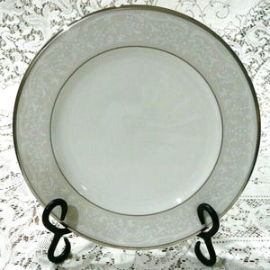 Może przedstawiać: Biały porcelanowy talerz z jasnoszarym kwiatowym obramowaniem i srebrną obwódką. Talerz jest prezentowany na czarnym metalowym stojaku, podkreślając jego elegancki design. Odpowiedni na formalne obiady.