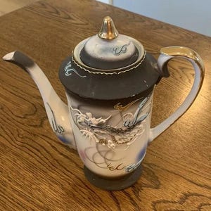 Peut inclure: Théière en céramique vintage avec un couvercle et une poignée noirs et dorés. Le corps de la théière est blanc avec un motif de dragon détaillé en relief. Le bec est noir et blanc, et la théière repose sur une surface en bois.