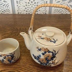 May include: Cream-colored ceramic teapot and teacup set. The teapot has a woven handle and is decorated with floral and branch designs in shades of blue and brown. The teacup has a matching design.