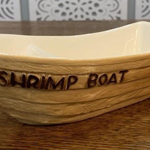Könnte beinhalten: Eine Keramikschale in Form eines kleinen Bootes mit hellbrauner Außenseite in Holzmaserungsoptik und dem Schriftzug SHRIMP BOAT in dunkelbraunen Buchstaben. Das Innere des Gefäßes ist glatt und mit einer glänzenden weißen Glasur versehen.