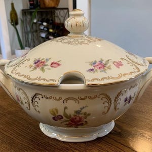 Pode incluir: Terrina de sopa de cerâmica creme com tampa, com detalhes florais e dourados. A terrina tem duas pegas e uma pequena abertura para uma concha. A tampa é encimada por um botão decorativo. Uma peça clássica para servir sopas e guisados.