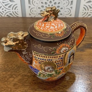 Op de afbeelding: Een decoratieve bruine theepot met een gouden drakensuitloop en handvat. Het deksel heeft een gouden drakenknop en oranje en witte patronen. De body is versierd met gedetailleerde illustraties en bloemmotieven.